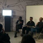 A picture of three black people sitting together with microphones in their hands in front of a small crowd