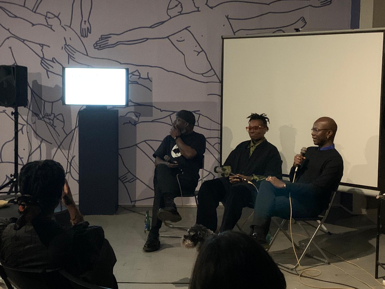 A picture of three black people sitting together with microphones in their hands in front of a small crowd