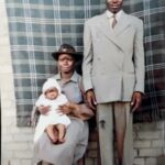 A picture of Black couple posing for a portrait. The woman holds a baby on her lap